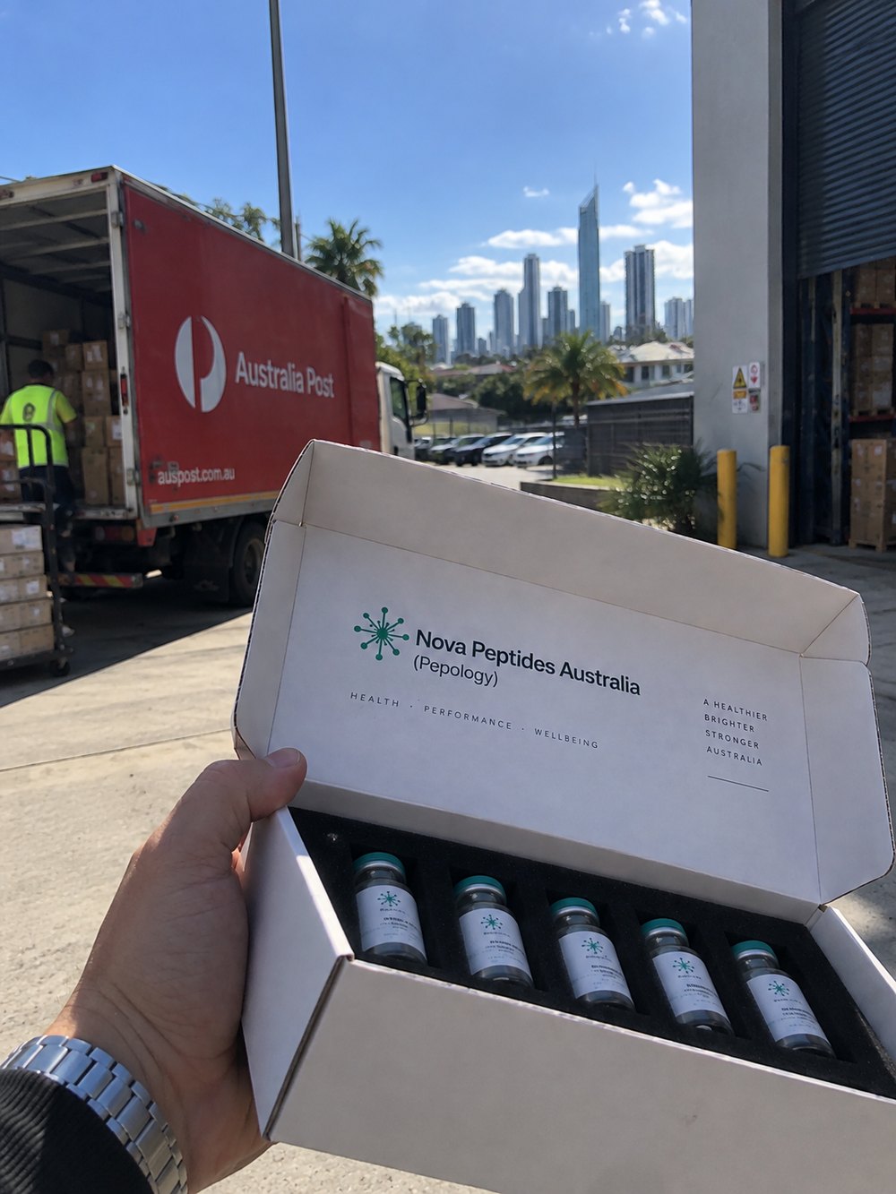A NovaPeptides Australia peptide kit held at the Gold Coast warehouse as an Australia Post truck is loaded for same-day dispatch, Surfers Paradise skyline behind